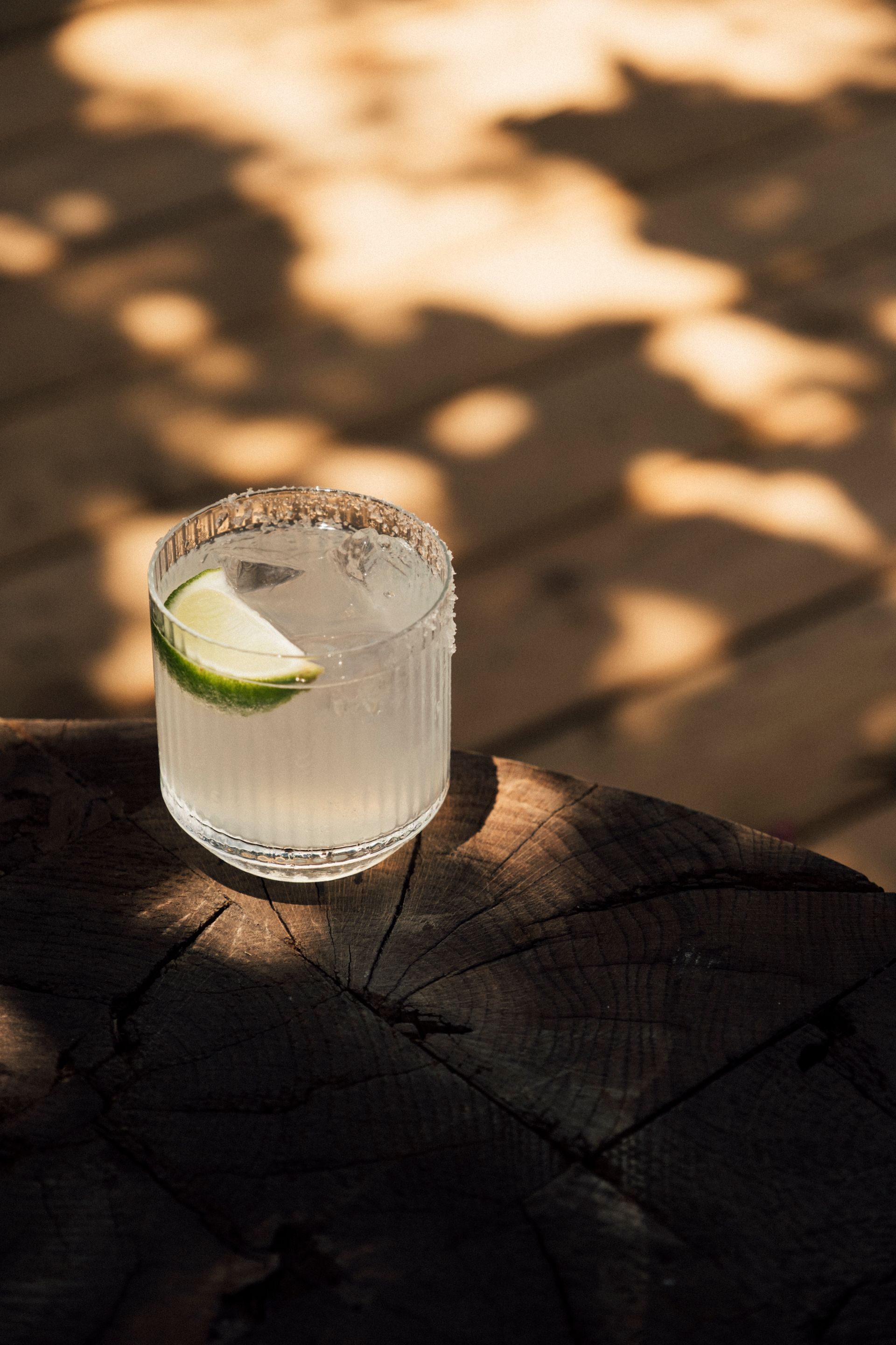 A glass of clear cocktail with a lime wedge garnish, sitting on a wooden surface with dappled sunlight.