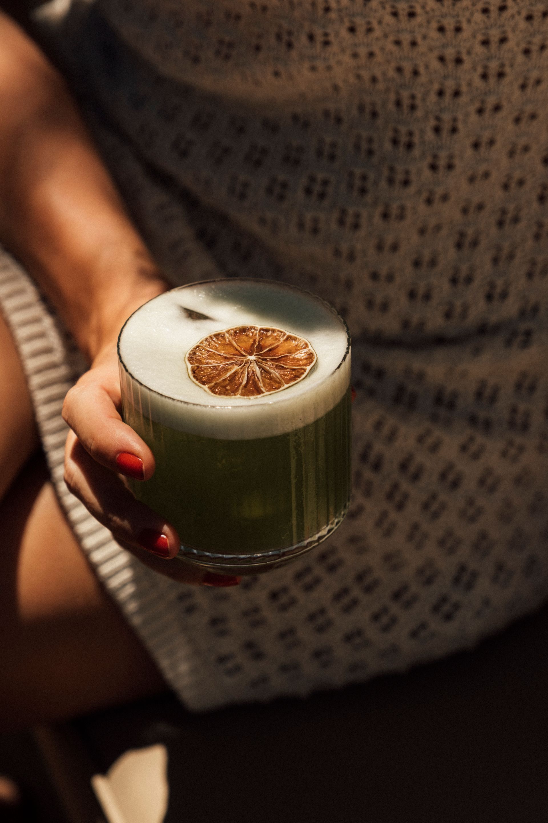 A person with red nail polish holds a chilled green cocktail in a textured glass, topped with a dehydrated citrus slice.