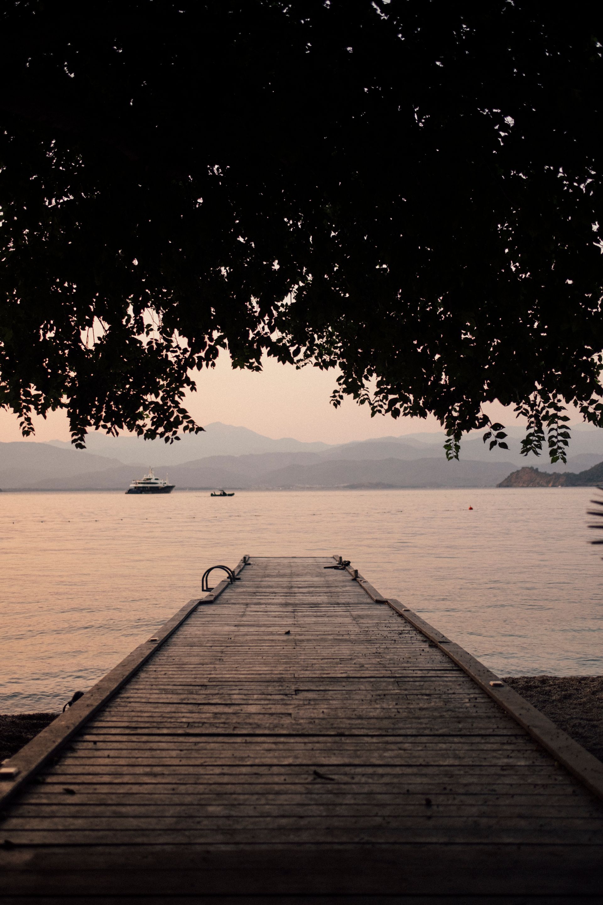 A wooden pier extends into a calm lake at sunset, framed by dark tree branches hanging overhead against distant mountains.