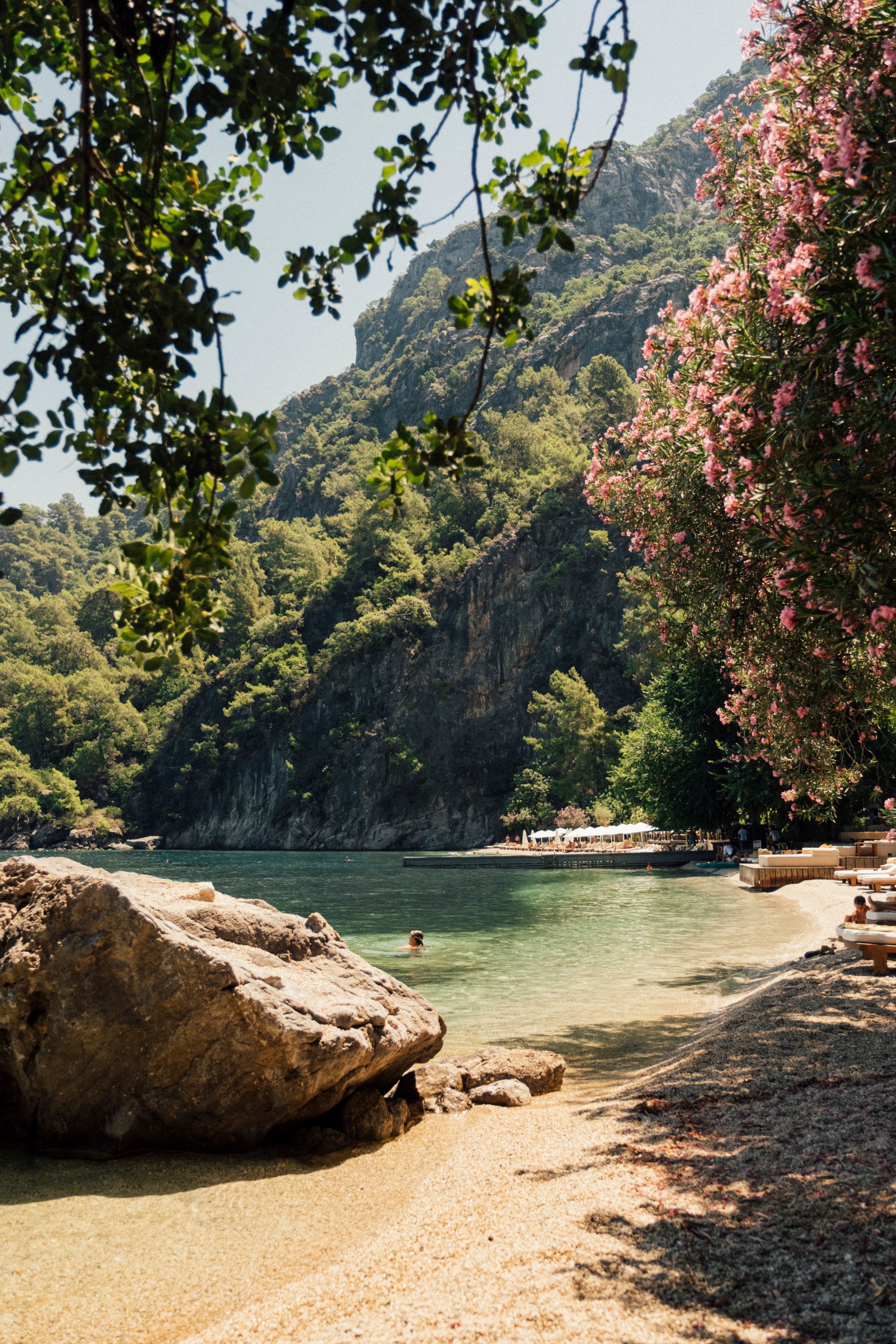 A pebbled beach with a large boulder in the foreground, calm turquoise water, and mountains covered in trees.