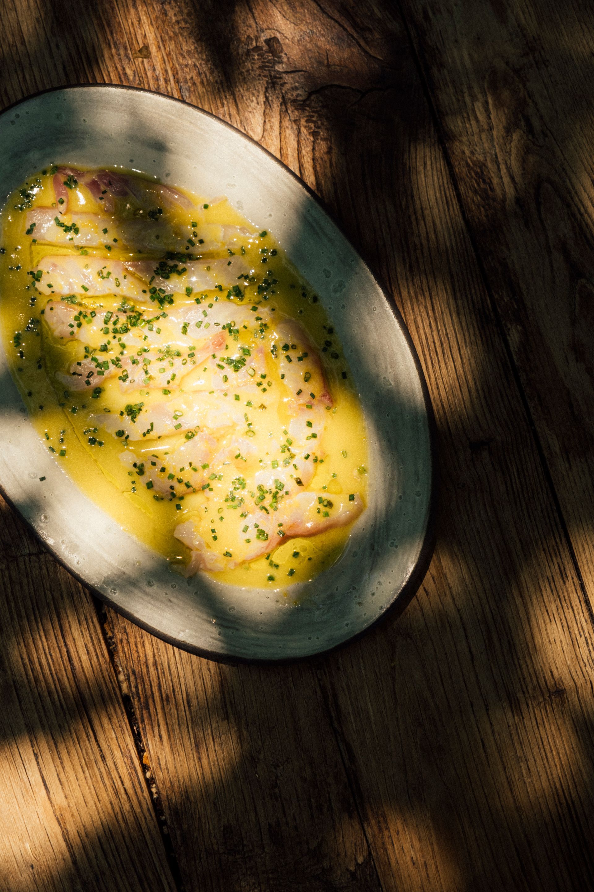 Thinly sliced raw fish carpaccio drizzled with oil and garnished with herbs, served on a plate on a rustic wooden table.