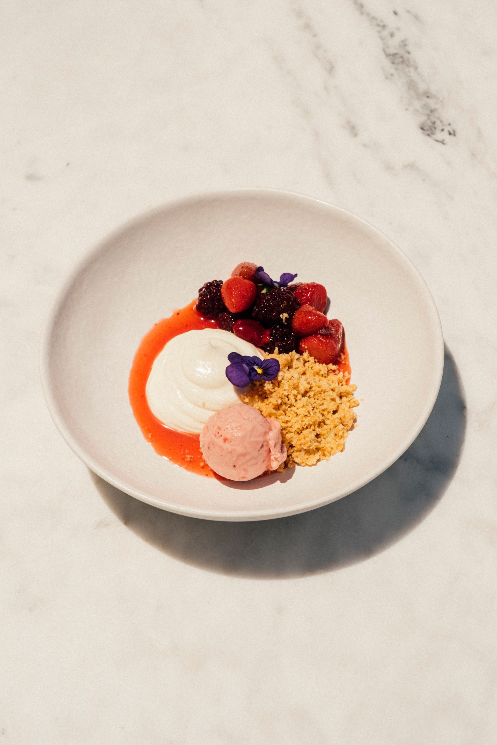 A gourmet dessert in a white bowl featuring fresh berries, a scoop of pink sorbet, cream, and crumble on a marble surface.