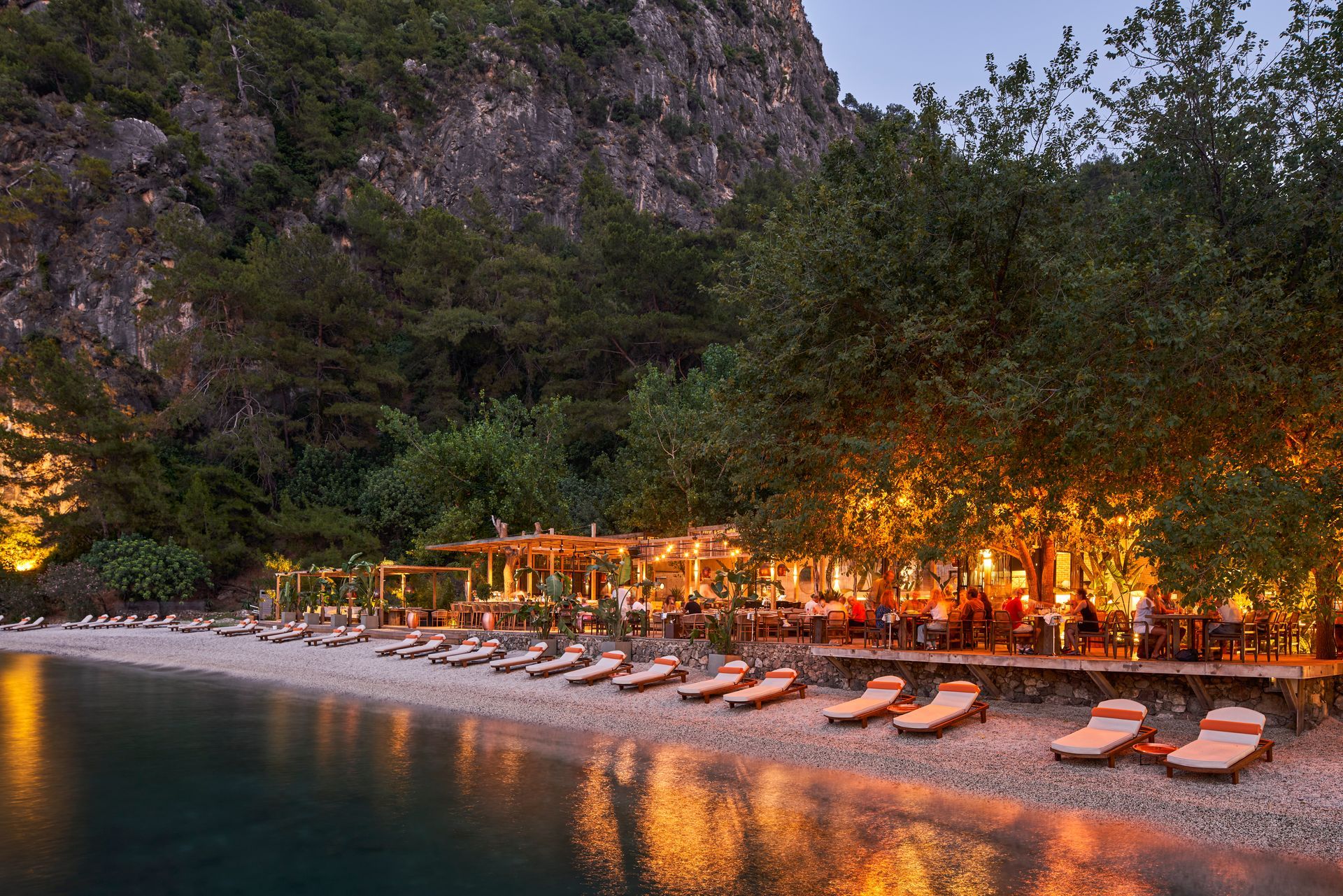 Lounge chairs line a pebble beach at twilight, with a restaurant illuminated by warm lights nestled under trees nearby.