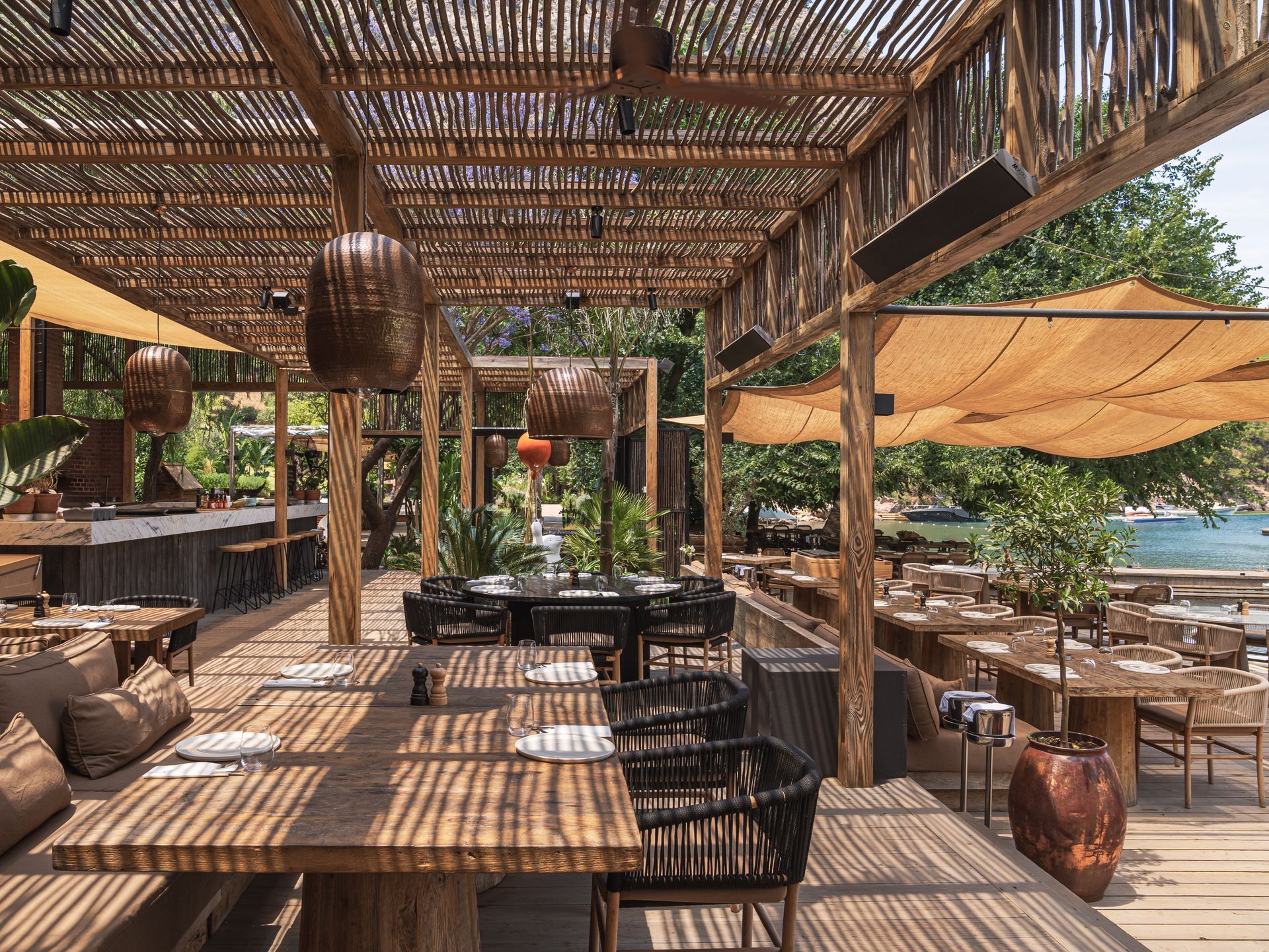 An outdoor restaurant terrace with wooden tables, woven shade roofs, wicker pendant lights, and a view of the water.