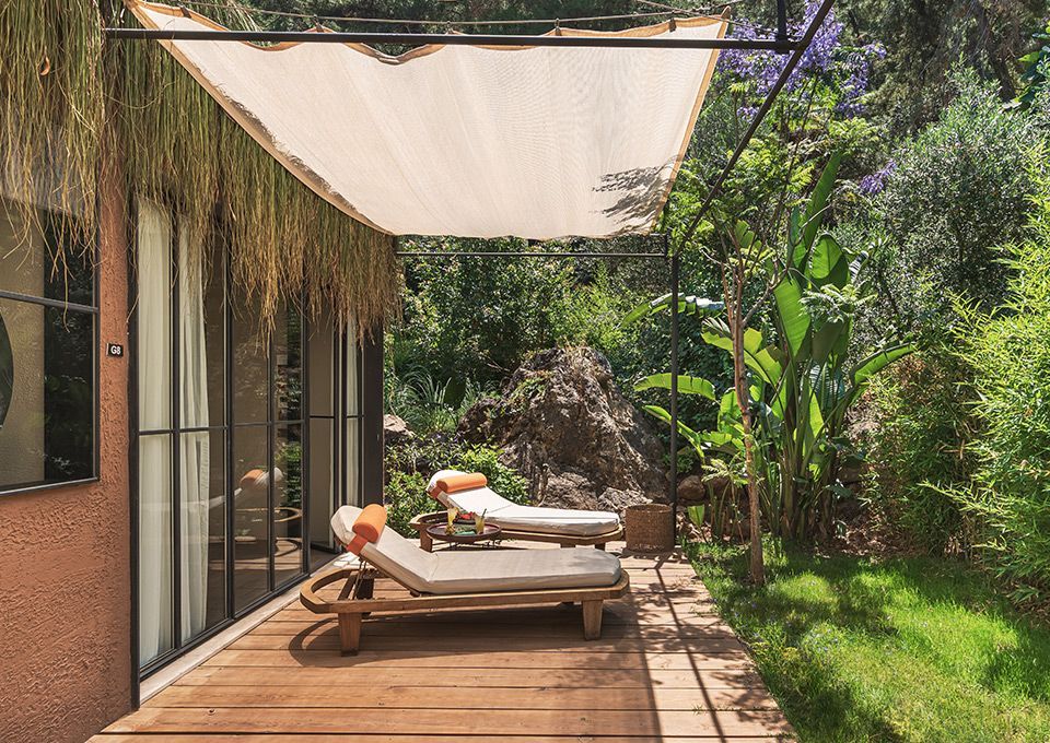 Two lounge chairs sit on a wooden deck under a shade sail, next to a building with glass doors and lush garden greenery.