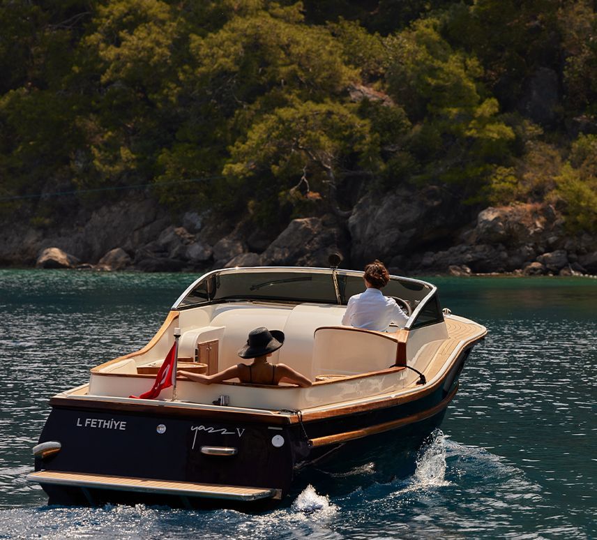 A classic dark blue and wood-trimmed motorboat with two passengers cruises through calm, turquoise coastal waters.