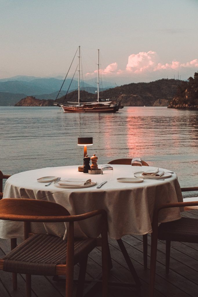 A candlelit dinner table set for two on a deck overlooking the ocean at sunset with a sailboat in the distance.