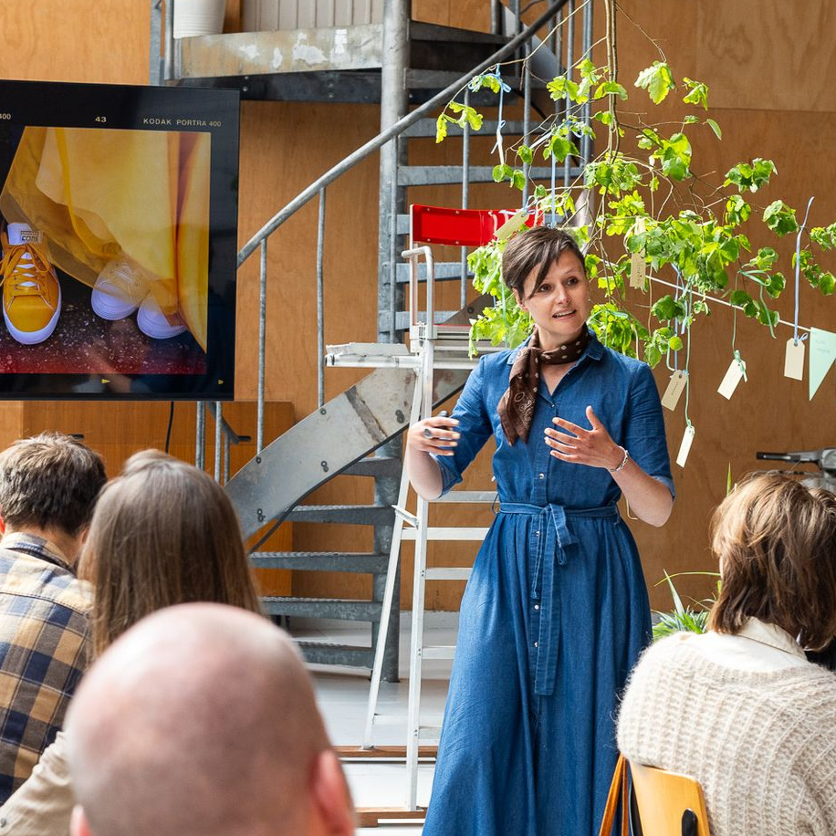 Myrna in een blauwe jurk spreekt voor een trap en een scherm, met een publiek dat in de buurt zit.