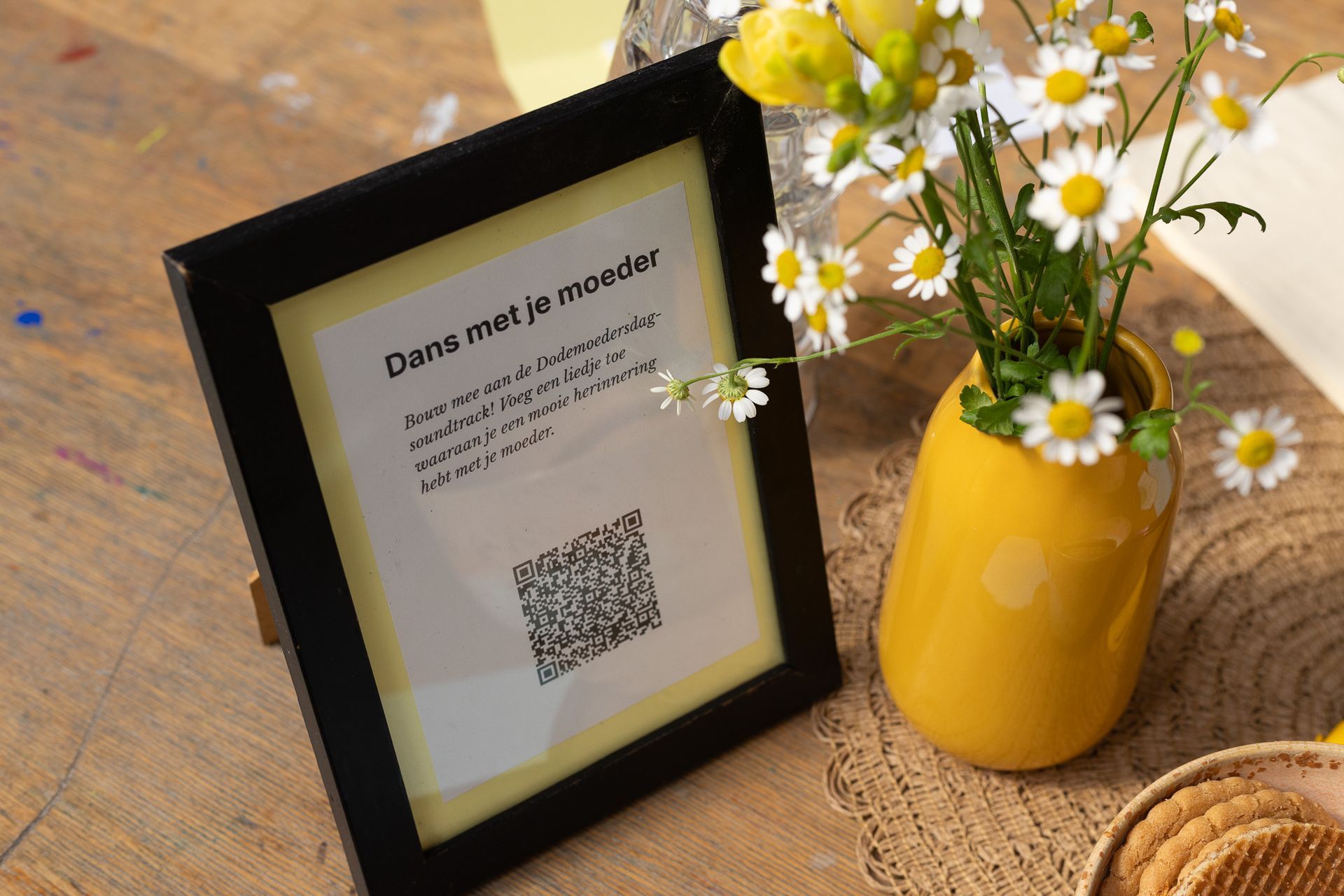 Ingelijst QR-codebordje naast een gele vaas met wilde bloemen op een rustieke houten tafel.