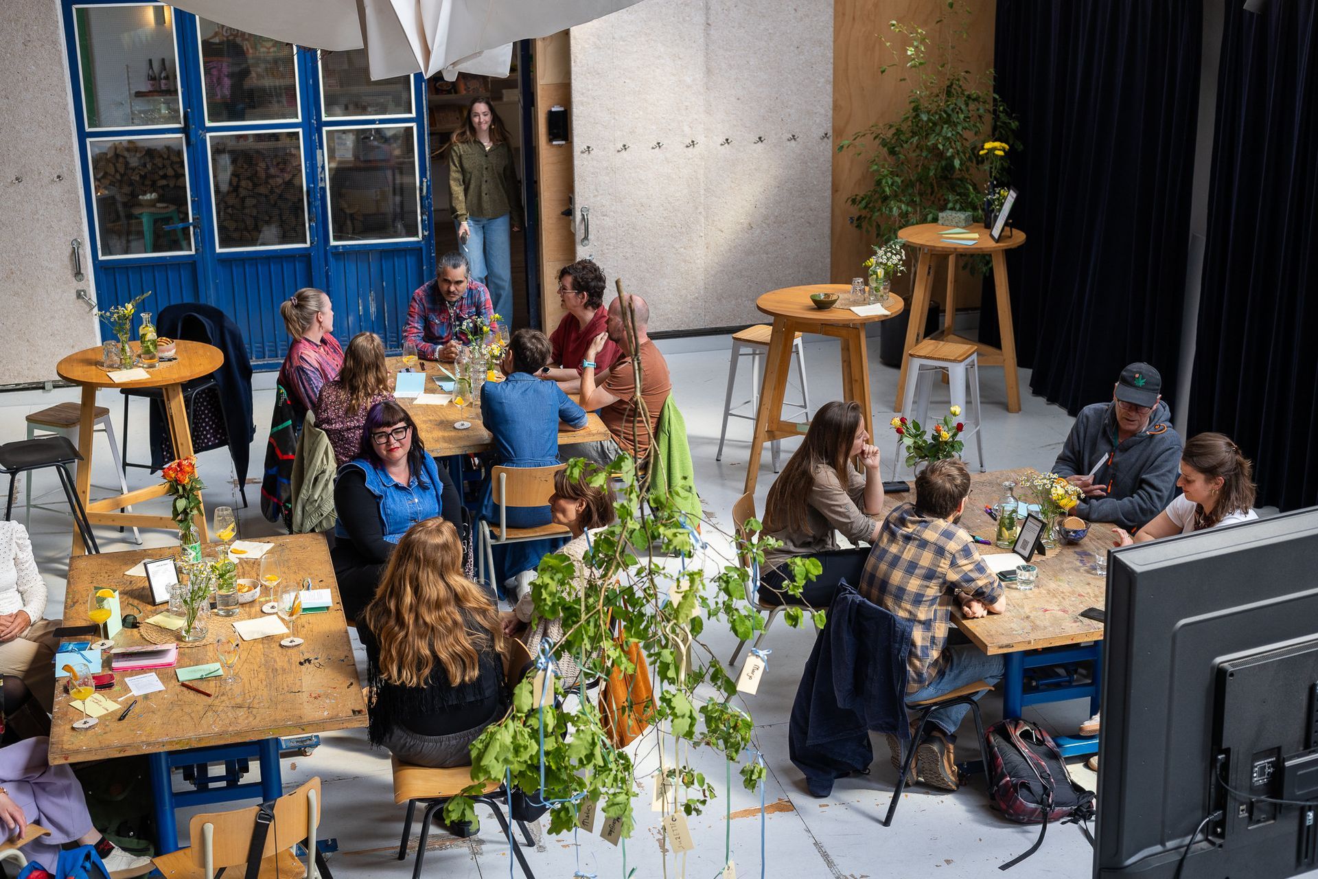 Mensen zitten rond tafels in een lichte ruimte, waar ze te midden van planten praatten met elkaar.