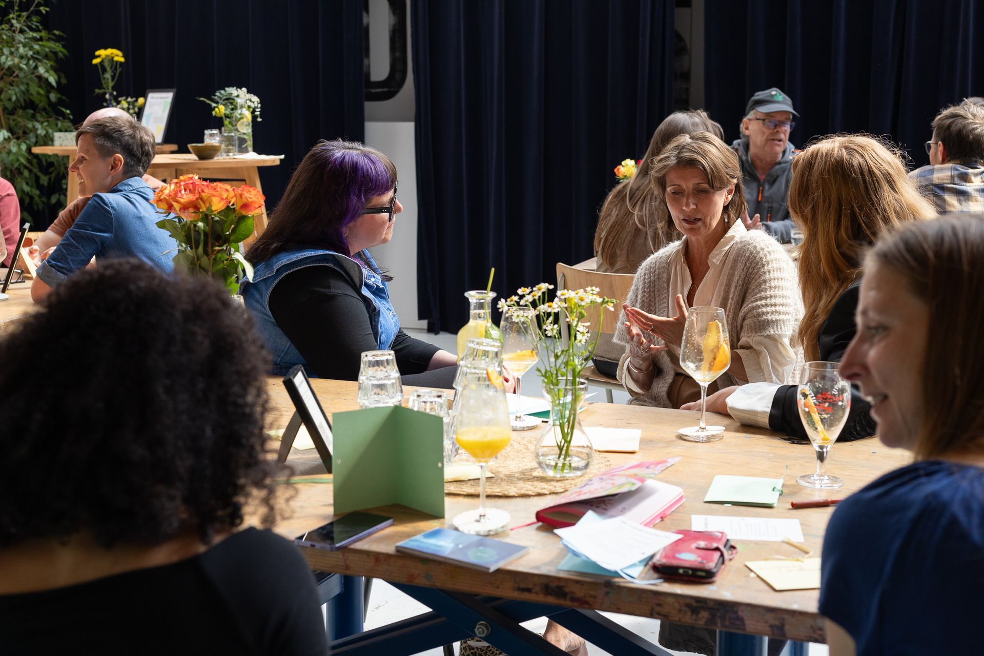 Mensen zitten rond een tafel met papieren, drankjes en bloemen tijdens Dodemoedersdag.