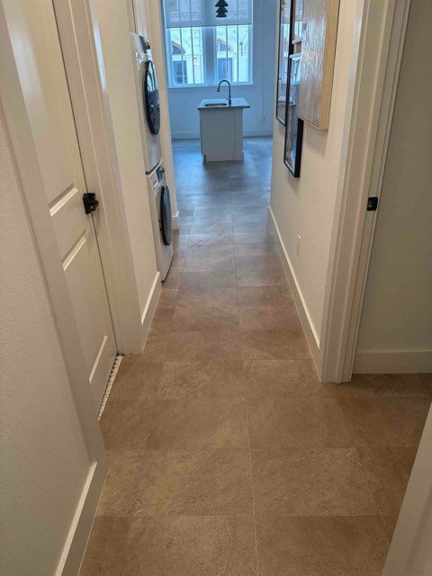 Hallway with light brown tiled floor, white trim and doors, leading to a kitchen with an island.