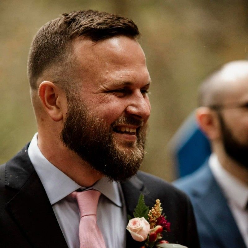 Man in a suit smiles, wearing a pink tie, with a dark beard.