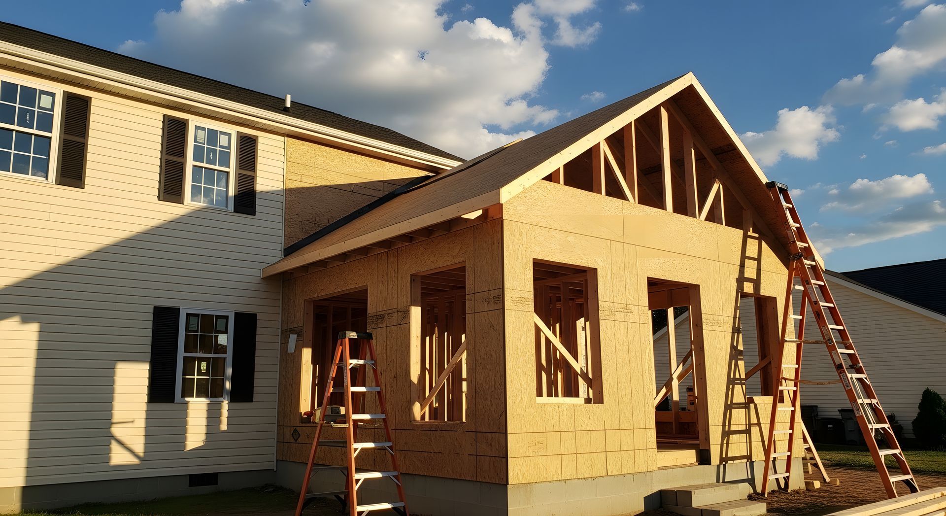 House under construction with exposed wood framing, ladders, and white siding in warm sunlight