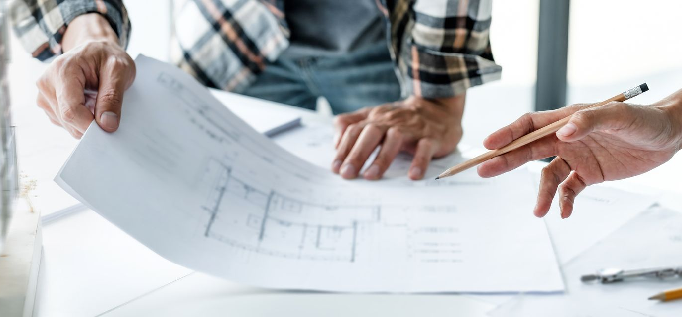 Two people reviewing architectural blueprints on a table; one points with a pencil.