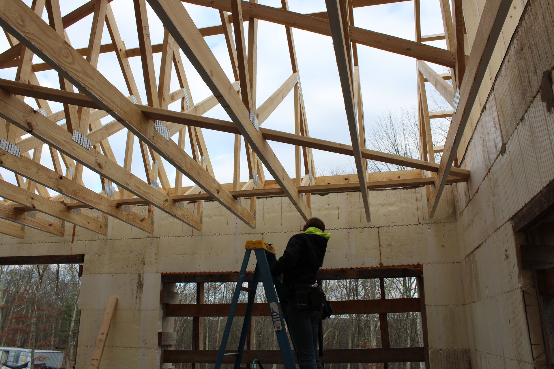 Construction worker on a ladder installing roof trusses in a building with wooden frames and a partly completed wall.