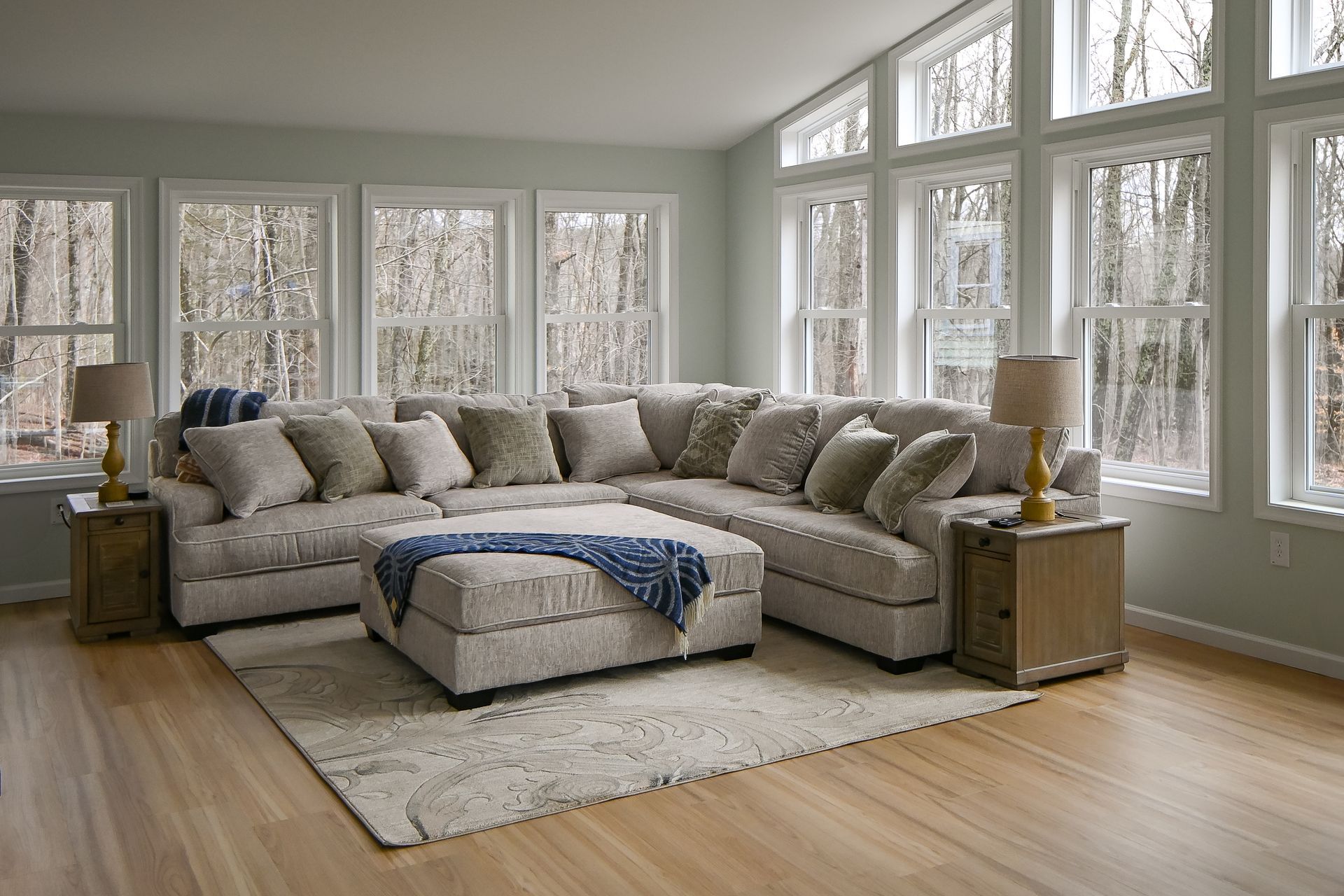 Light-filled living room with a sectional sofa and ottoman, surrounded by windows. Beige and neutral tones.