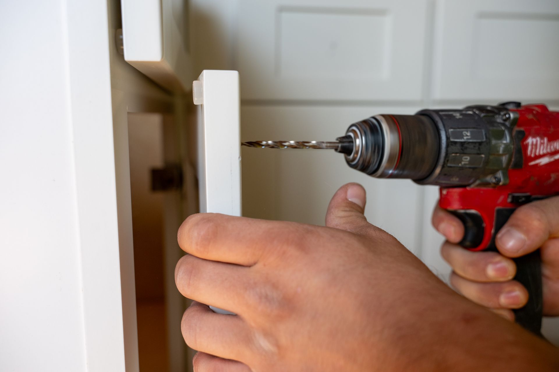Close-up of a hand holding a red power drill, boring a hole into a white cabinet door.