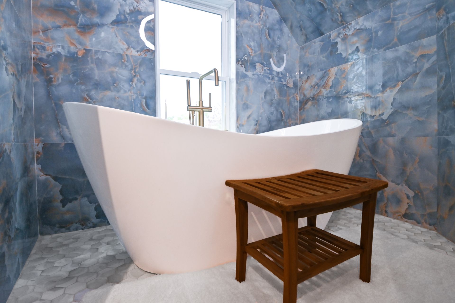White freestanding bathtub in a modern bathroom with blue-toned tile and a wooden stool.