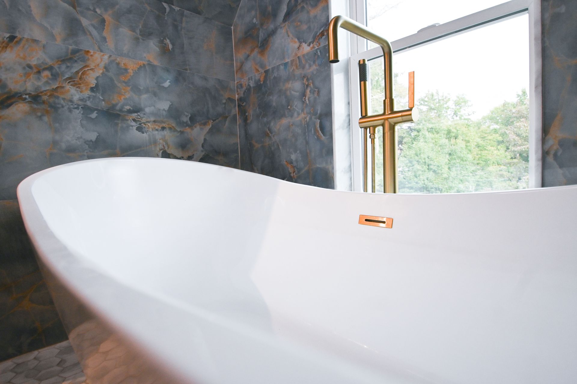 White oval bathtub with gold faucet next to a window and marble-like tile wall.