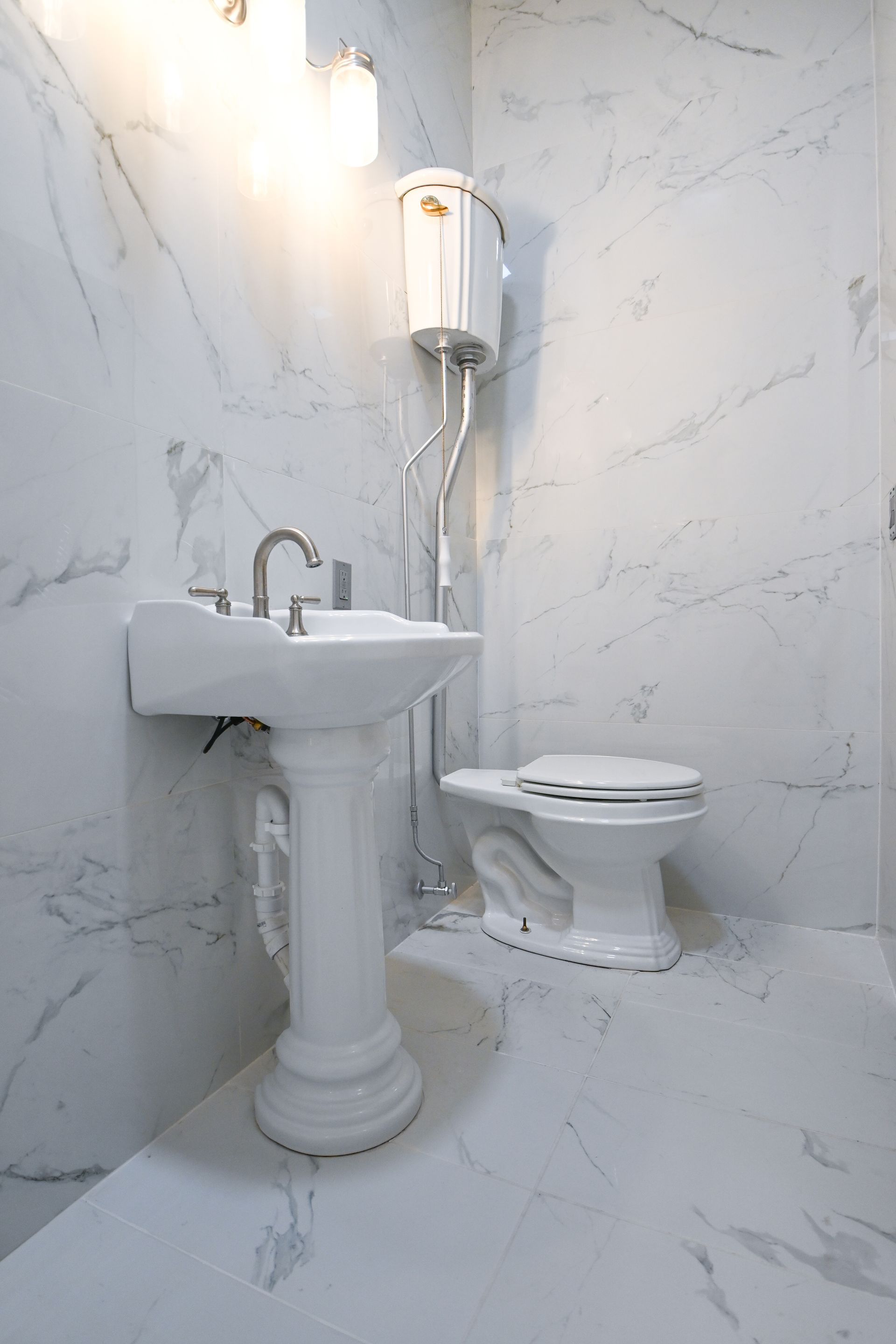 White marble bathroom with pedestal sink, toilet, and overhead water tank.