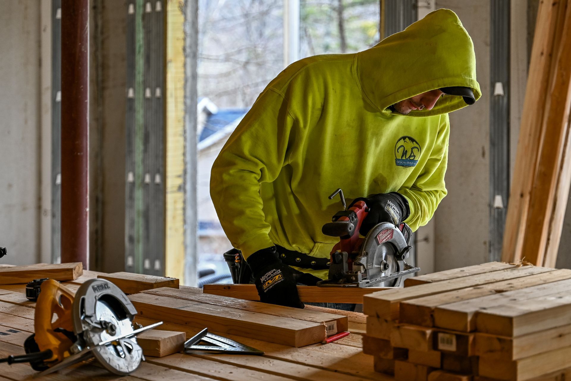 Person in yellow hoodie uses a circular saw on wood. Indoors, construction site, sunlight.