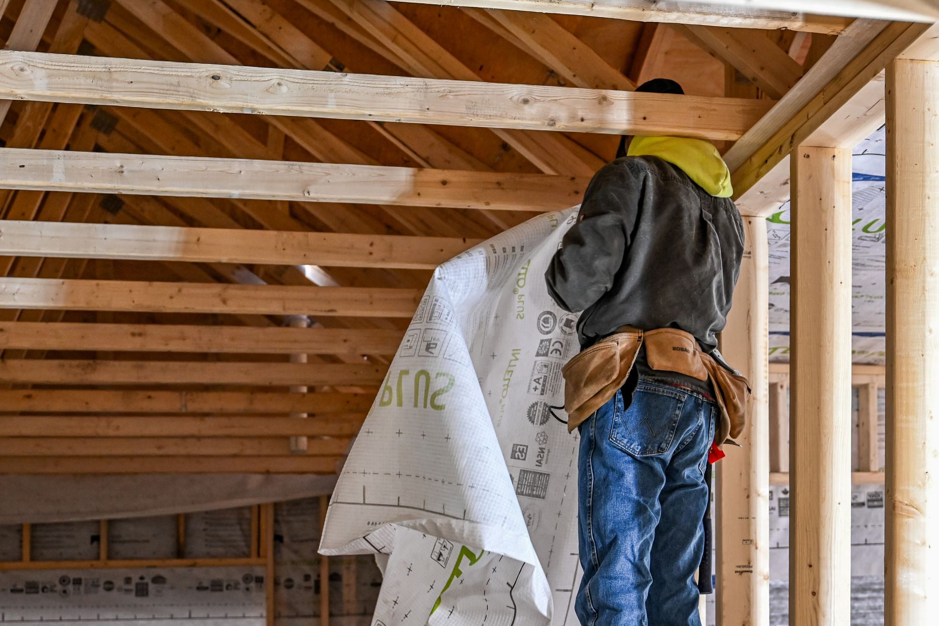 Worker in yellow hard hat and hoodie installing insulation in an unfinished wooden attic framing