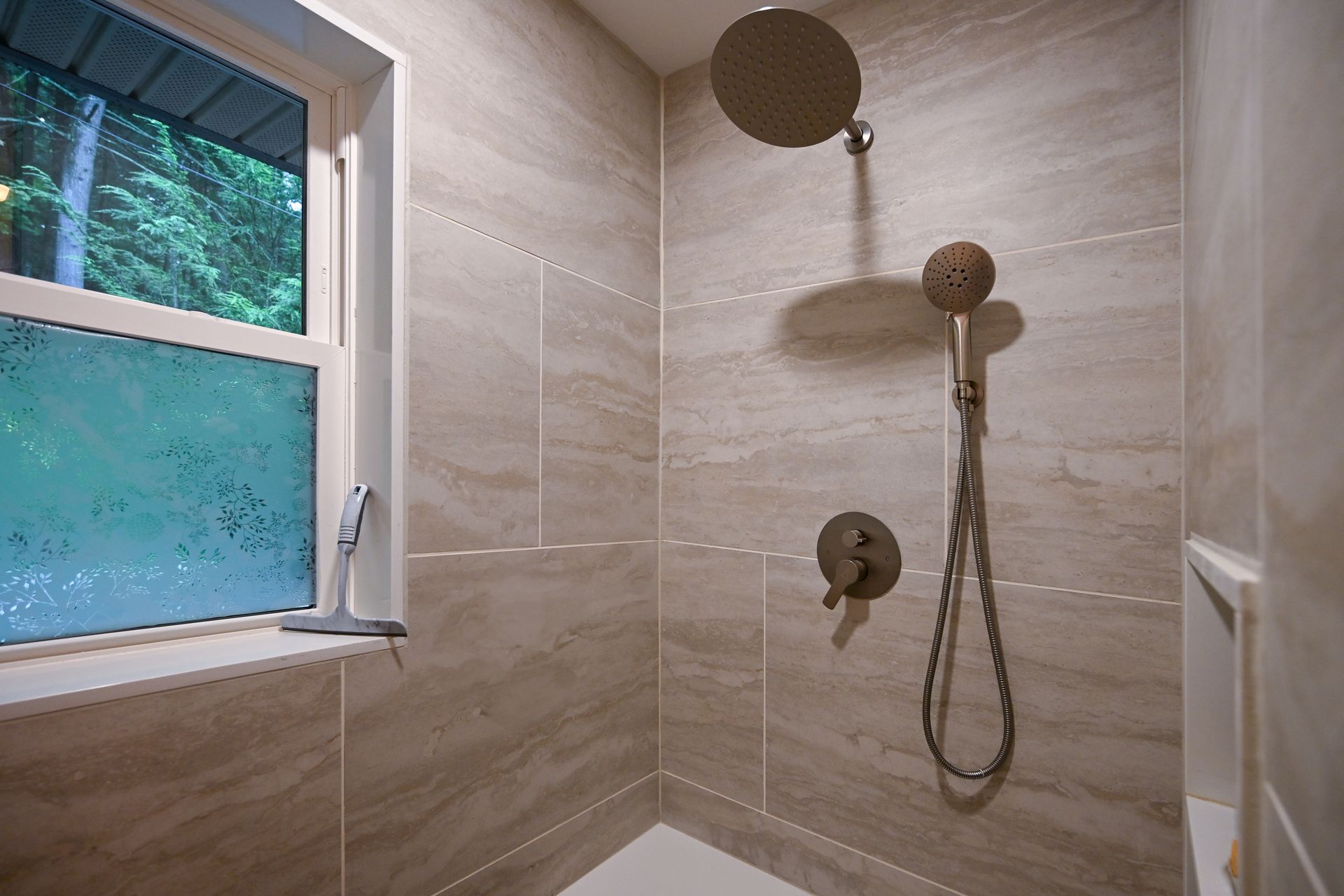 Modern shower with tan tiles, rain shower head, and window overlooking trees. Modern shower with tan tiles, rain shower head, and window overlooking trees.