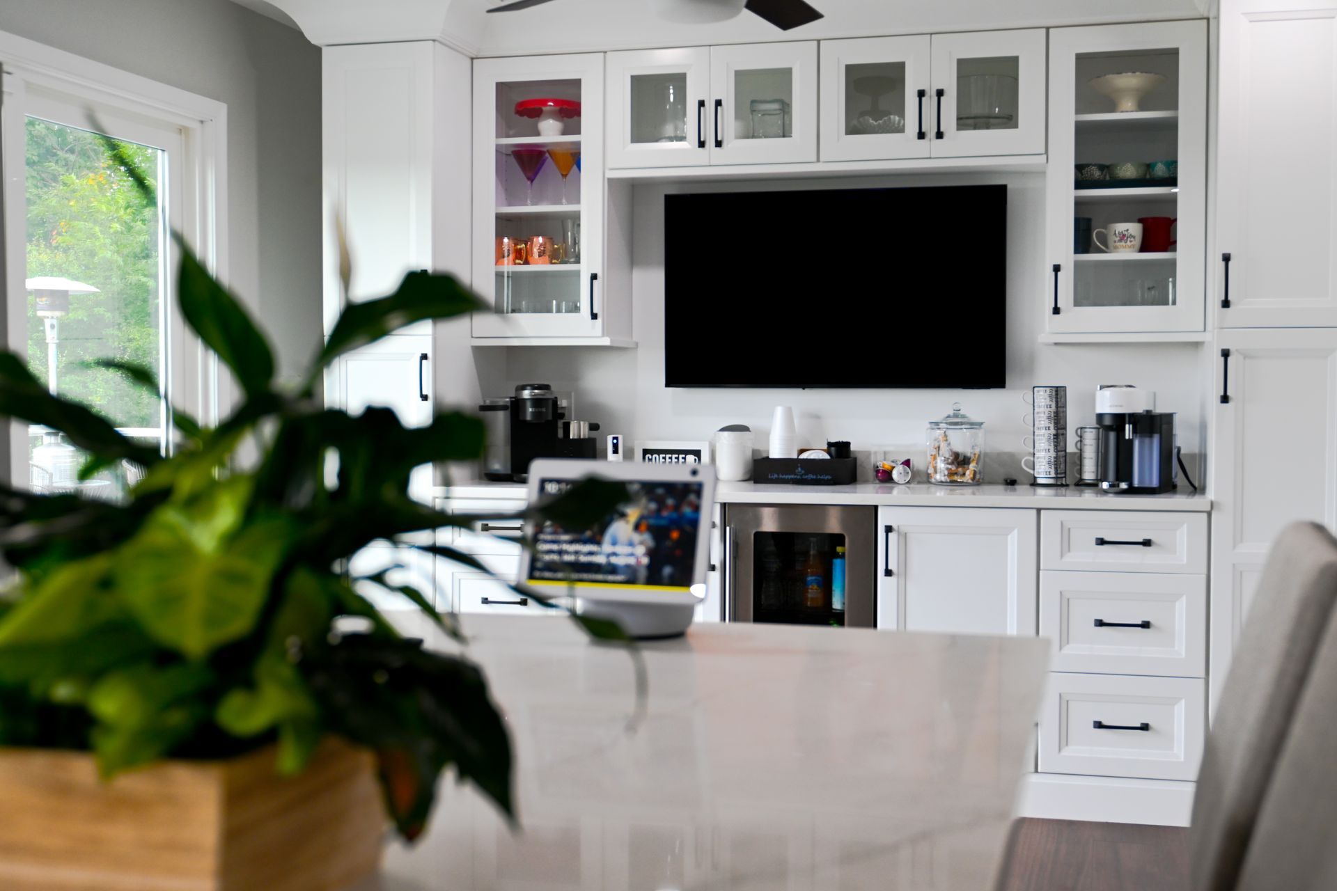 Kitchen with white cabinets, built-in TV, and countertop appliances. Plant in foreground, window on left. Kitchen with white cabinets, built-in TV, and countertop appliances. Plant in foreground, window on left.