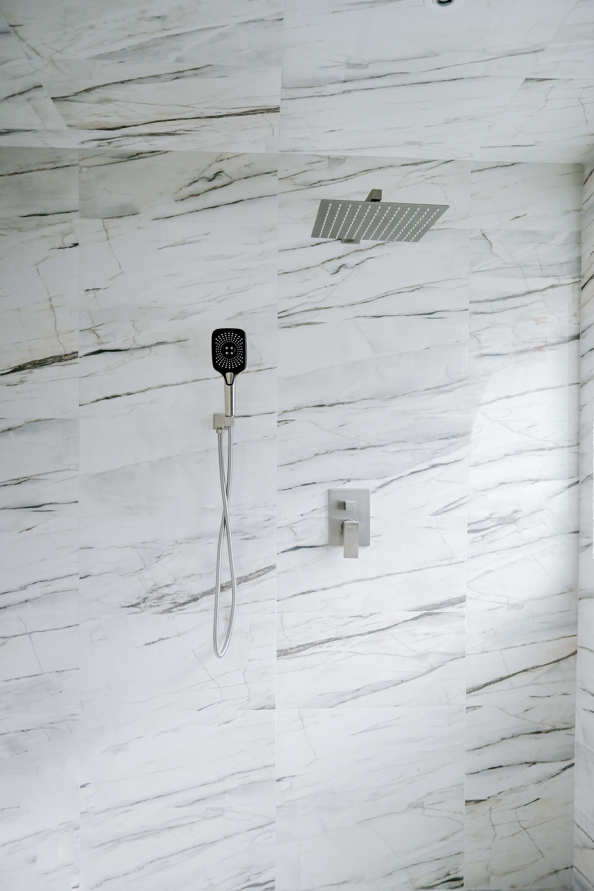 Shower with marble-patterned white tiles, square rain showerhead, handheld shower, and control valve.