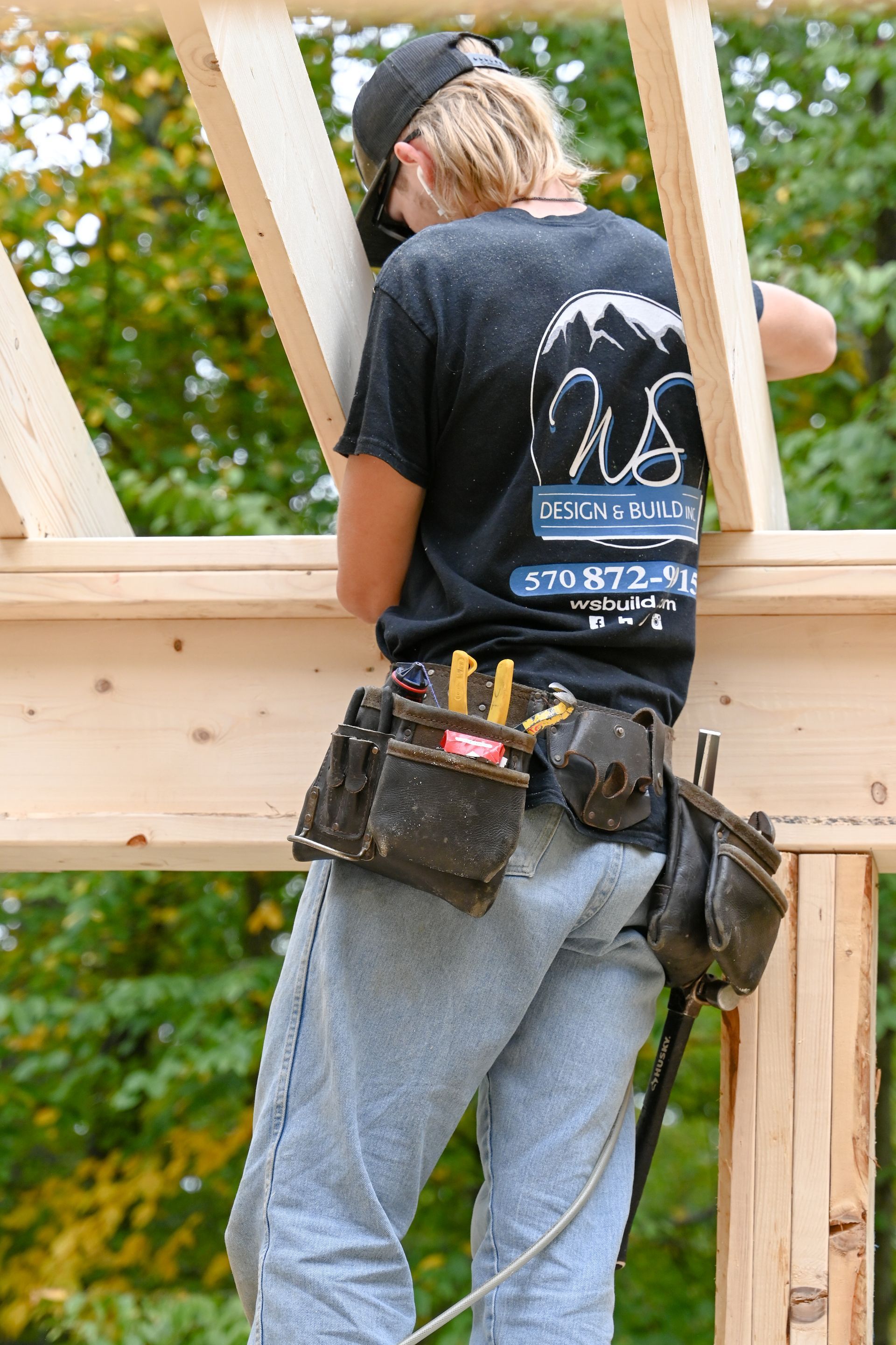 WS Design & Build worker with tool belt, wearing a black shirt, inspecting wooden beams.