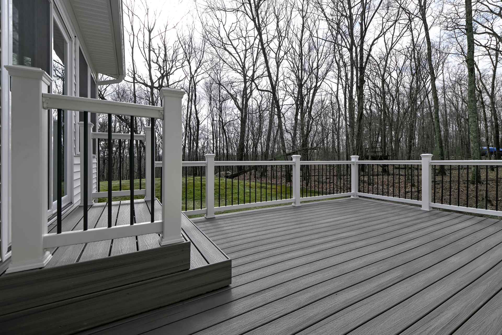 Gray composite deck with white railing, black balusters, and woods in the background.