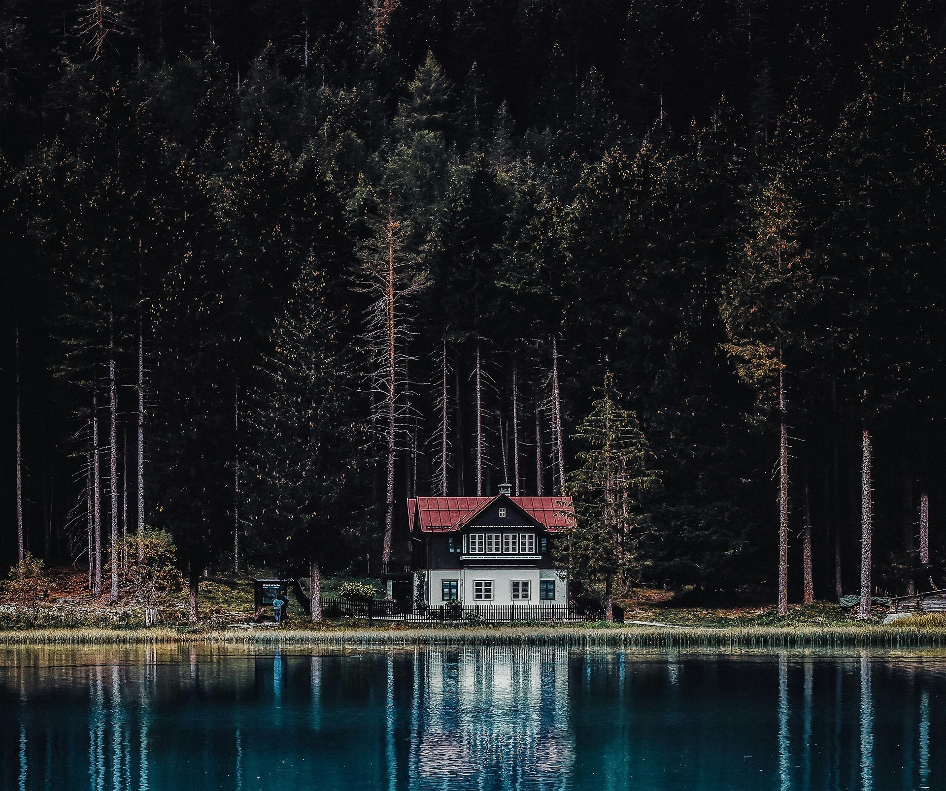 House with a red roof reflected in calm blue lake, nestled among dark evergreen trees.