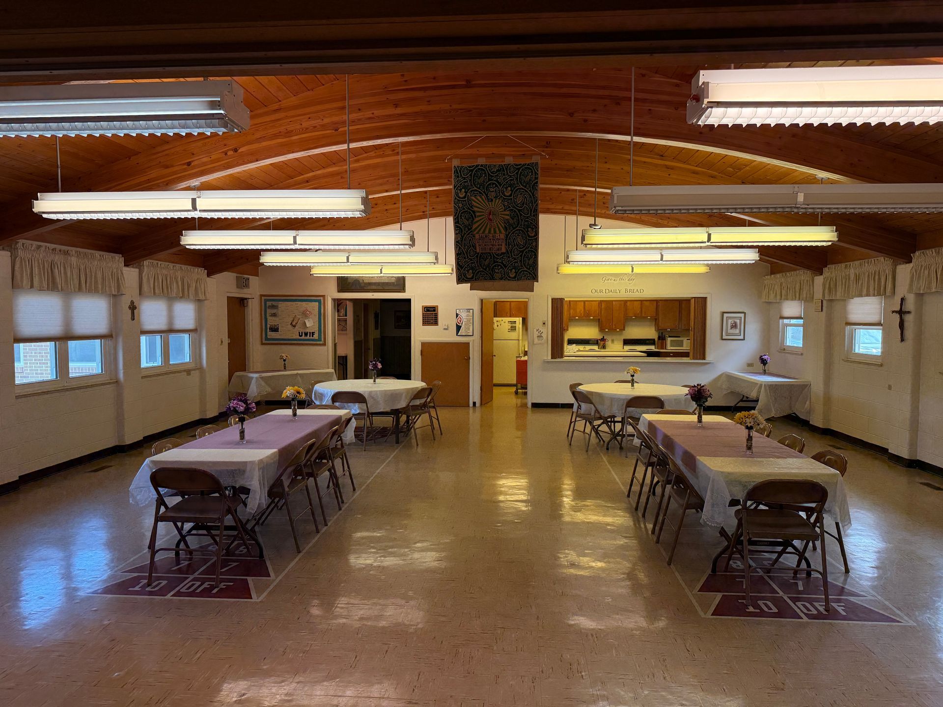 Community hall with rows of folding tables and chairs under fluorescent lights and a wood ceiling.