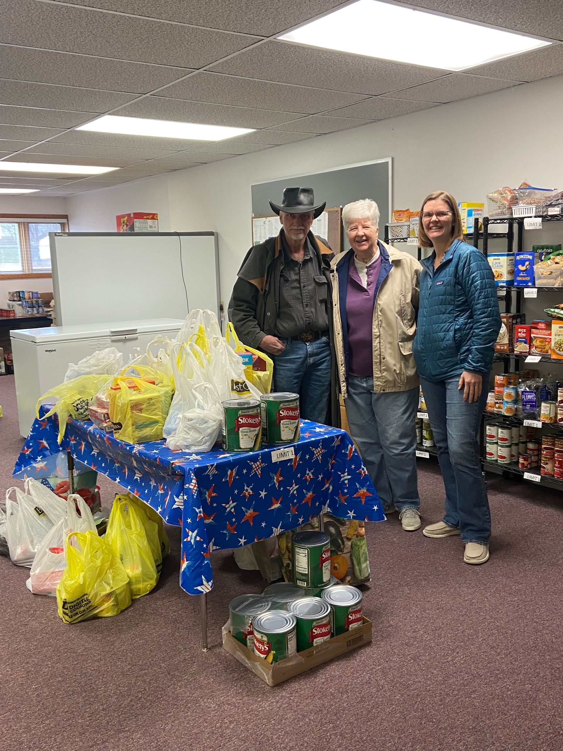 Three people stand behind a table laden with food donations in a small store, smiling.
