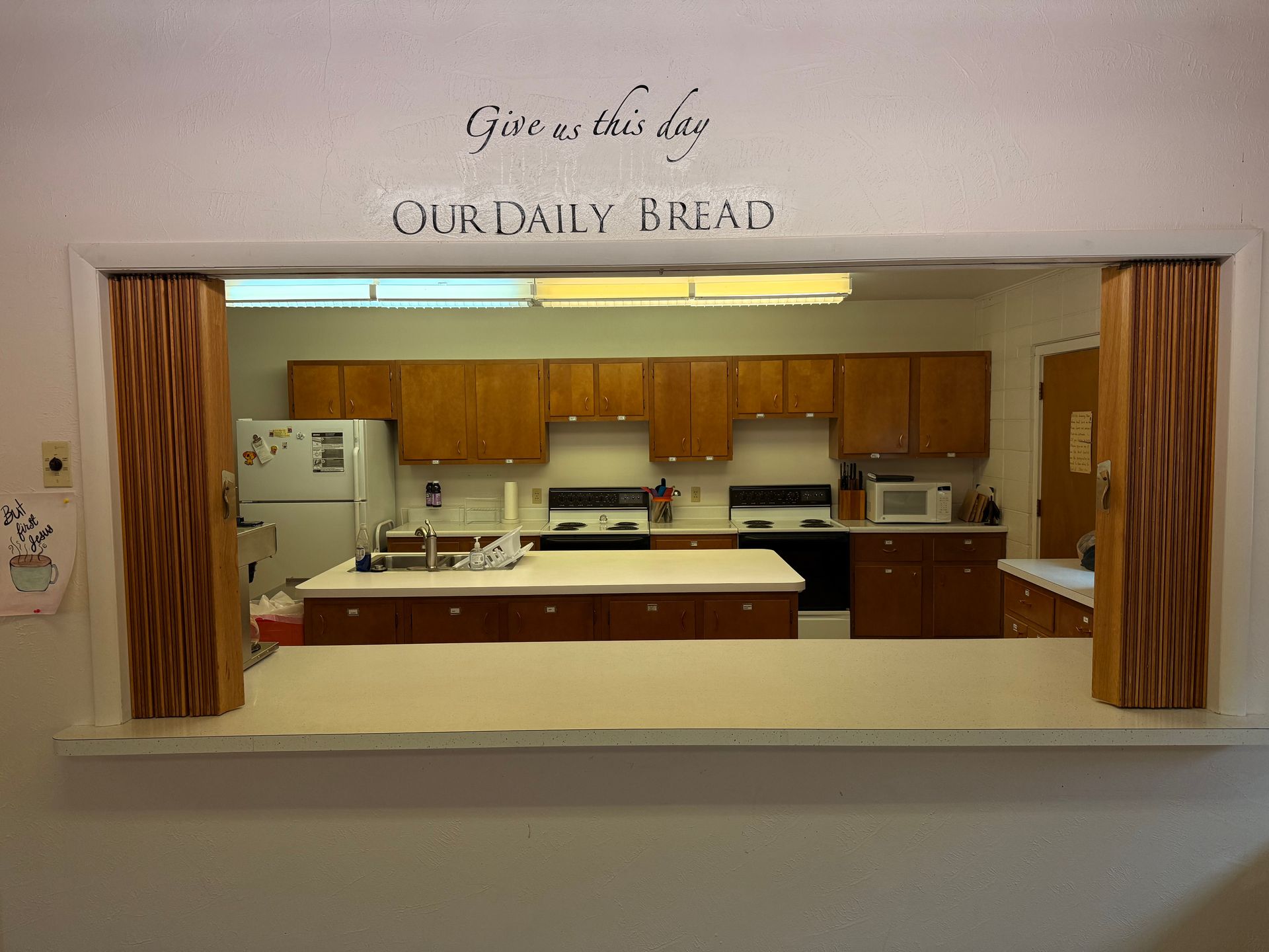 Framed view of a kitchen with wood cabinets and white appliances, labeled “Our Daily Bread” above the opening
