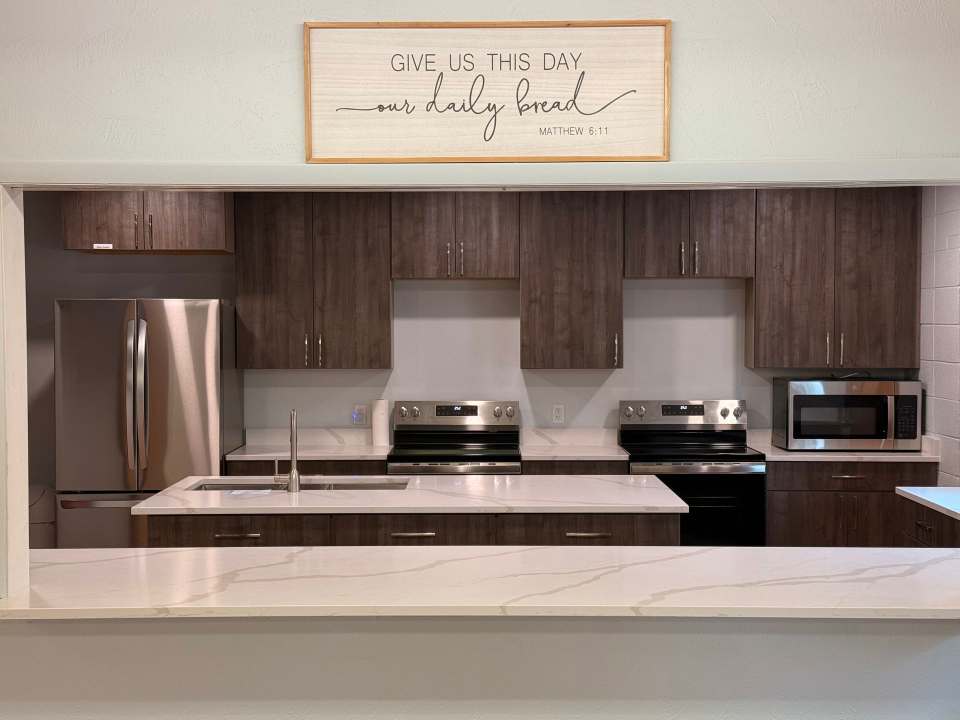 Modern kitchen with stainless steel appliances, dark wood cabinets, white counters, and a framed sign above the stove.