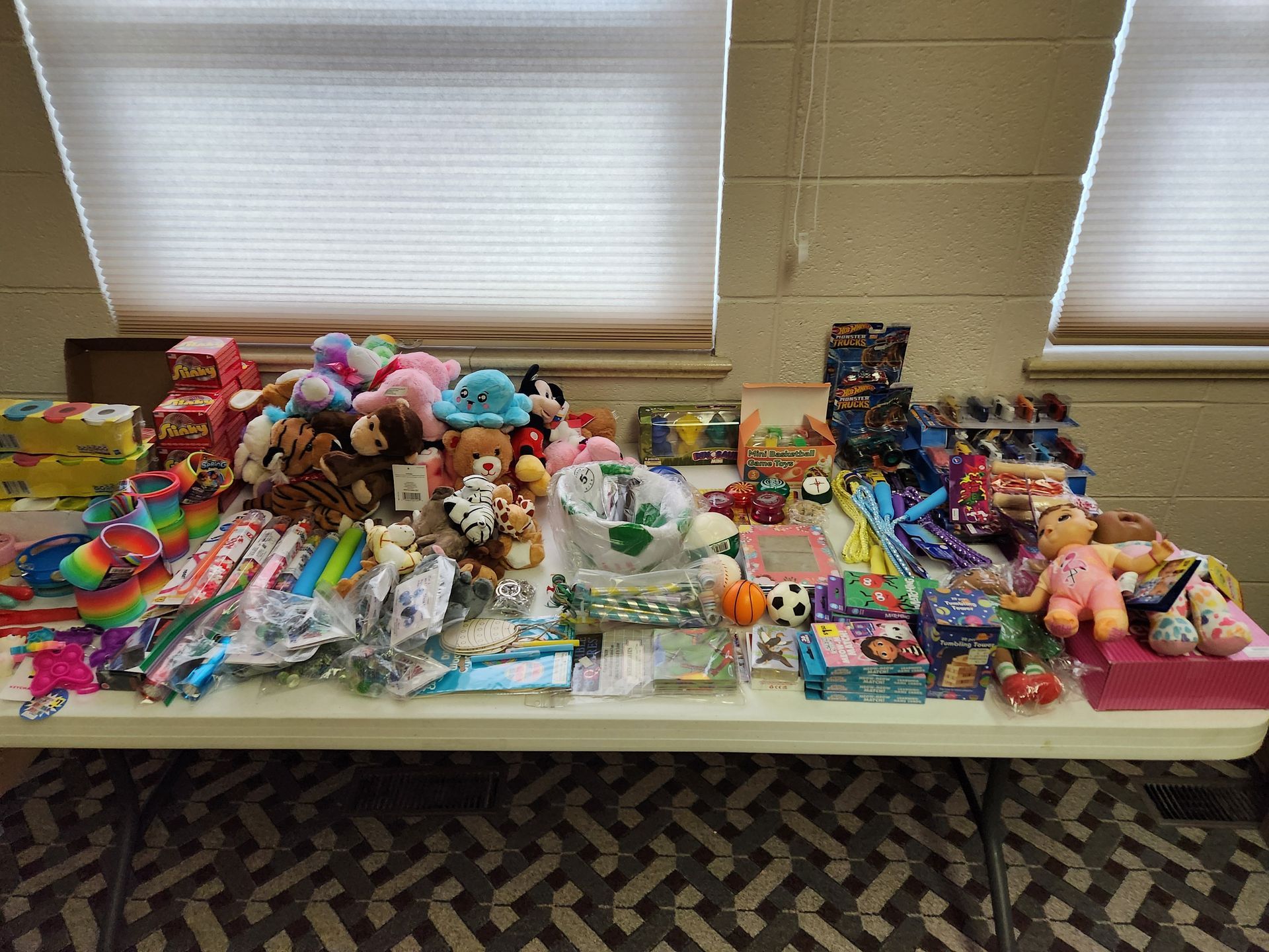 Table covered with assorted toys, dolls, candy, and colorful small items beneath two windows.