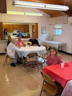 People at tables in a community hall, some talking and eating. Kitchen in background.