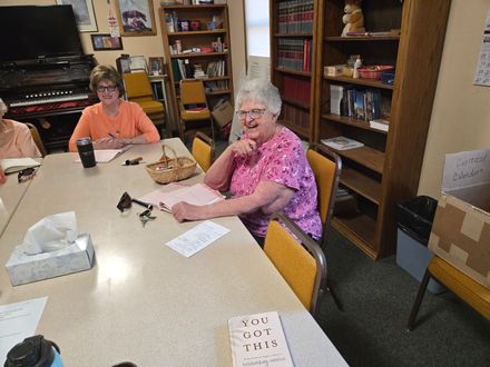 People seated around a table in a room with shelves and boxes, one woman smiling and writing on paper.