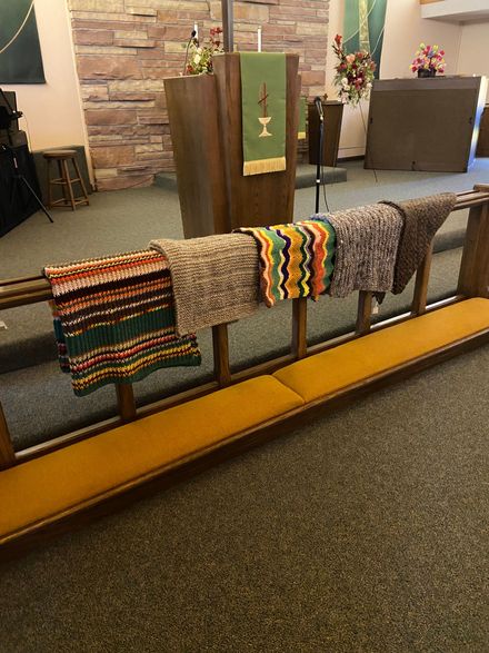 Colorful crocheted scarves draped over a metal rail in a church-like interior with stone walls.