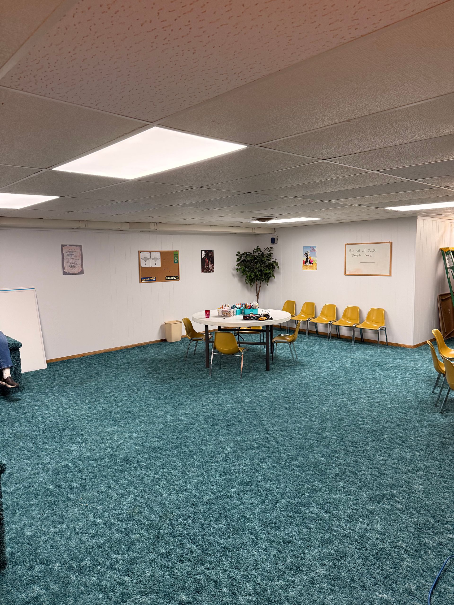 Empty classroom or meeting room with teal carpet, white walls, tables, and yellow chairs under fluorescent lights
