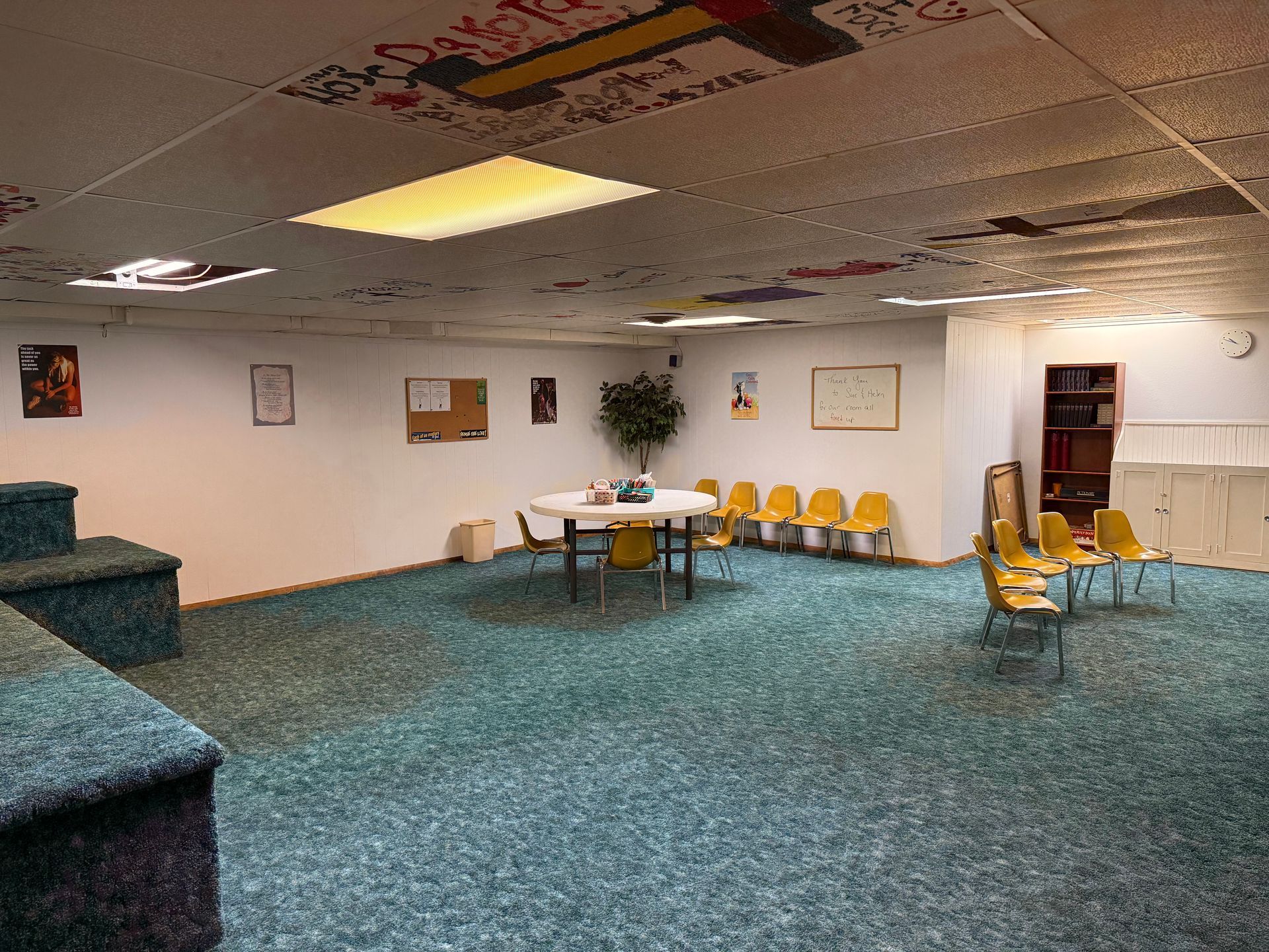 Carpeted room with white walls, tables, yellow chairs, and a small stage on the left.