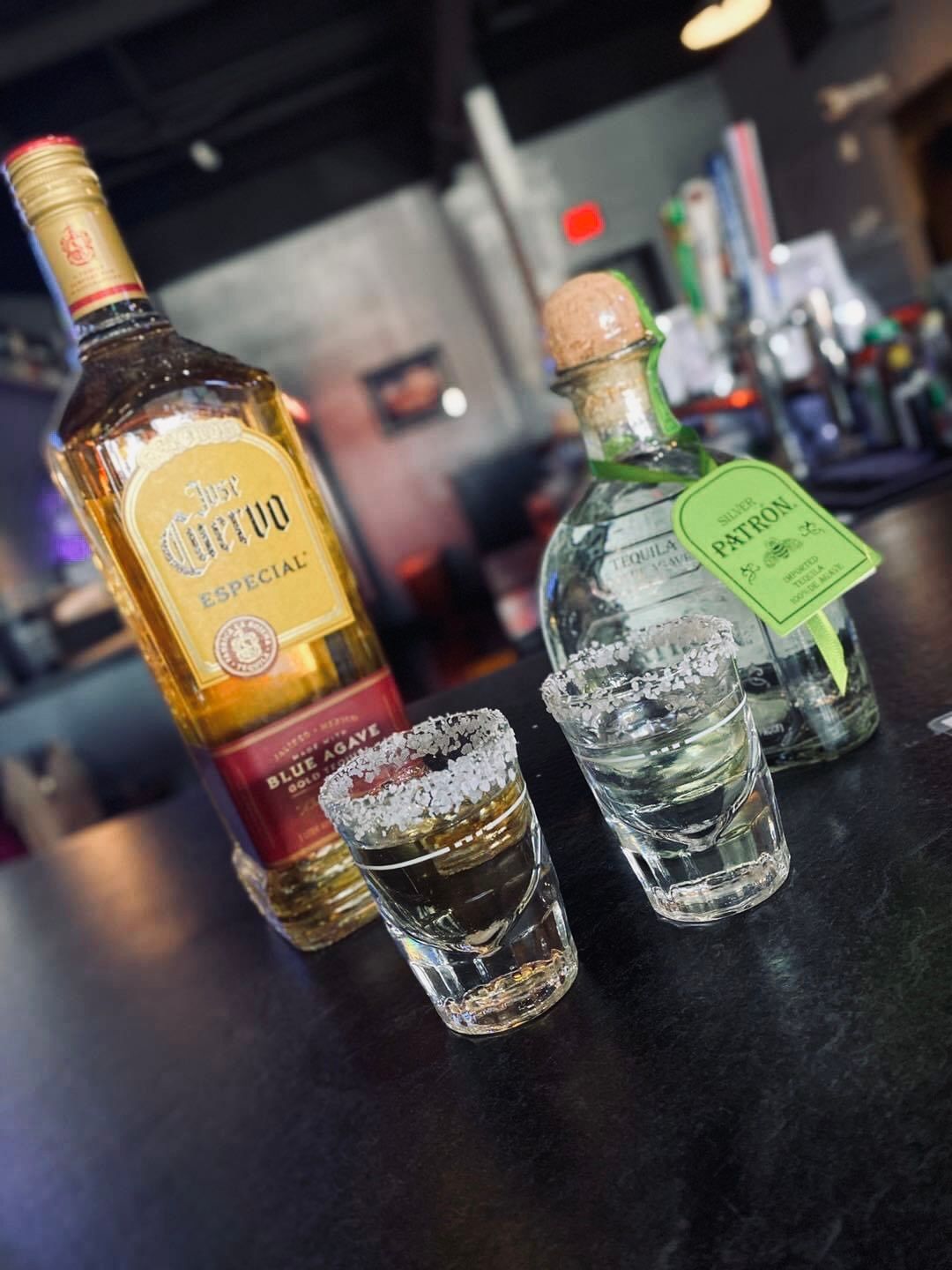 Tequila bottles with two shot glasses, one rimmed with salt, on a dark bar.