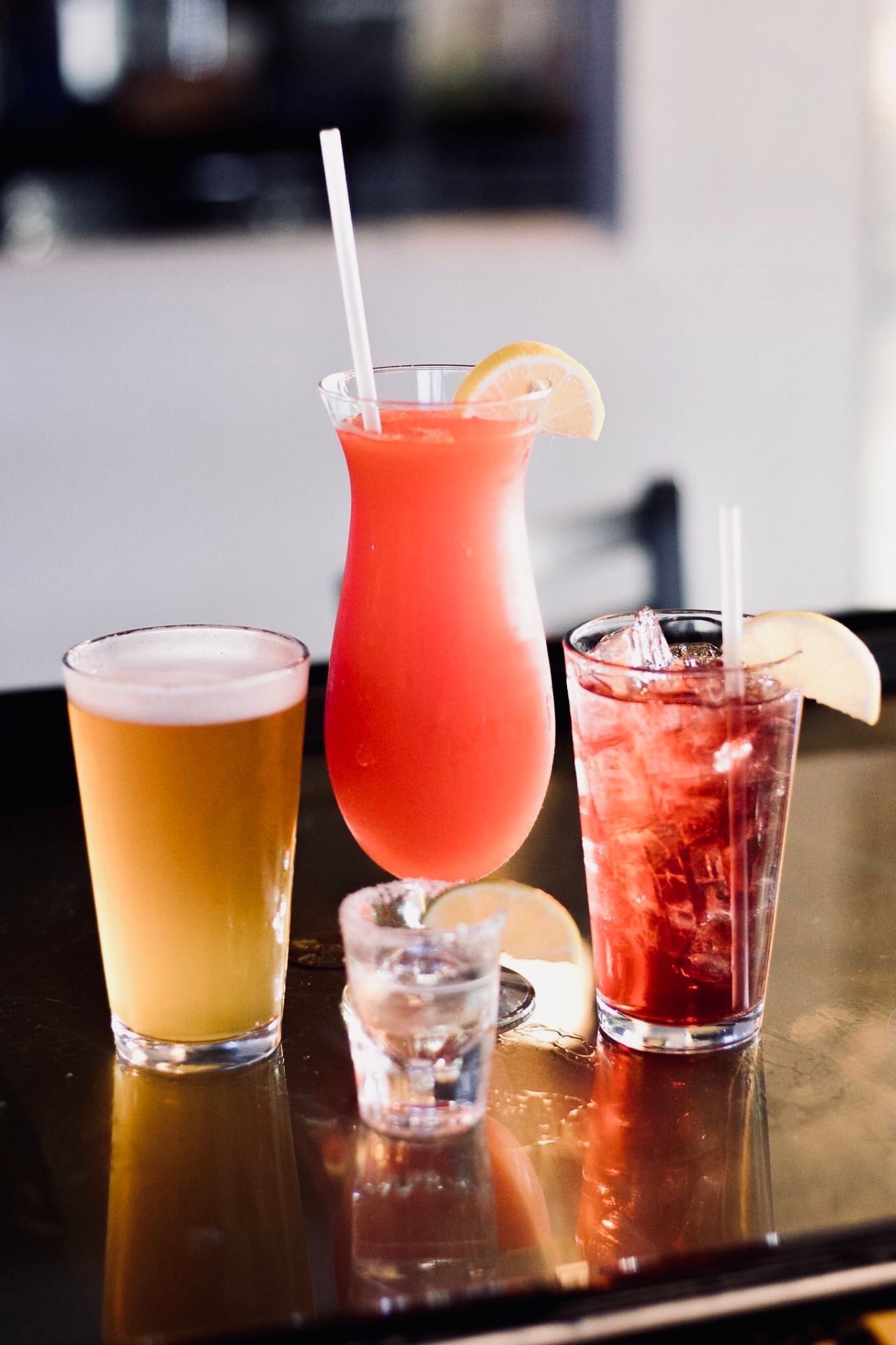Drinks on a bar: beer, red cocktail with straw, red cocktail with ice and lemon, and shot glass.