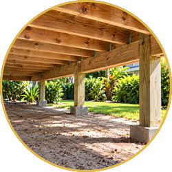 Wooden raised deck with support posts over a shaded gravel area beside lush green plants