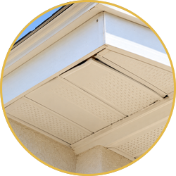 Underside of a house roof eave with white soffit vents and beige siding under blue sky