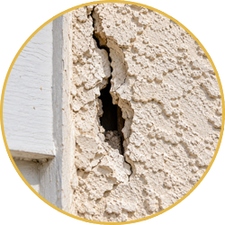 Close-up of a cracked stucco wall with a large vertical hole near a window trim