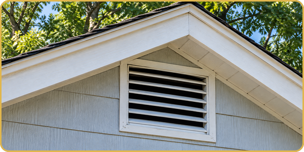 White house gable with a small louvered attic vent under the roofline and green trees above