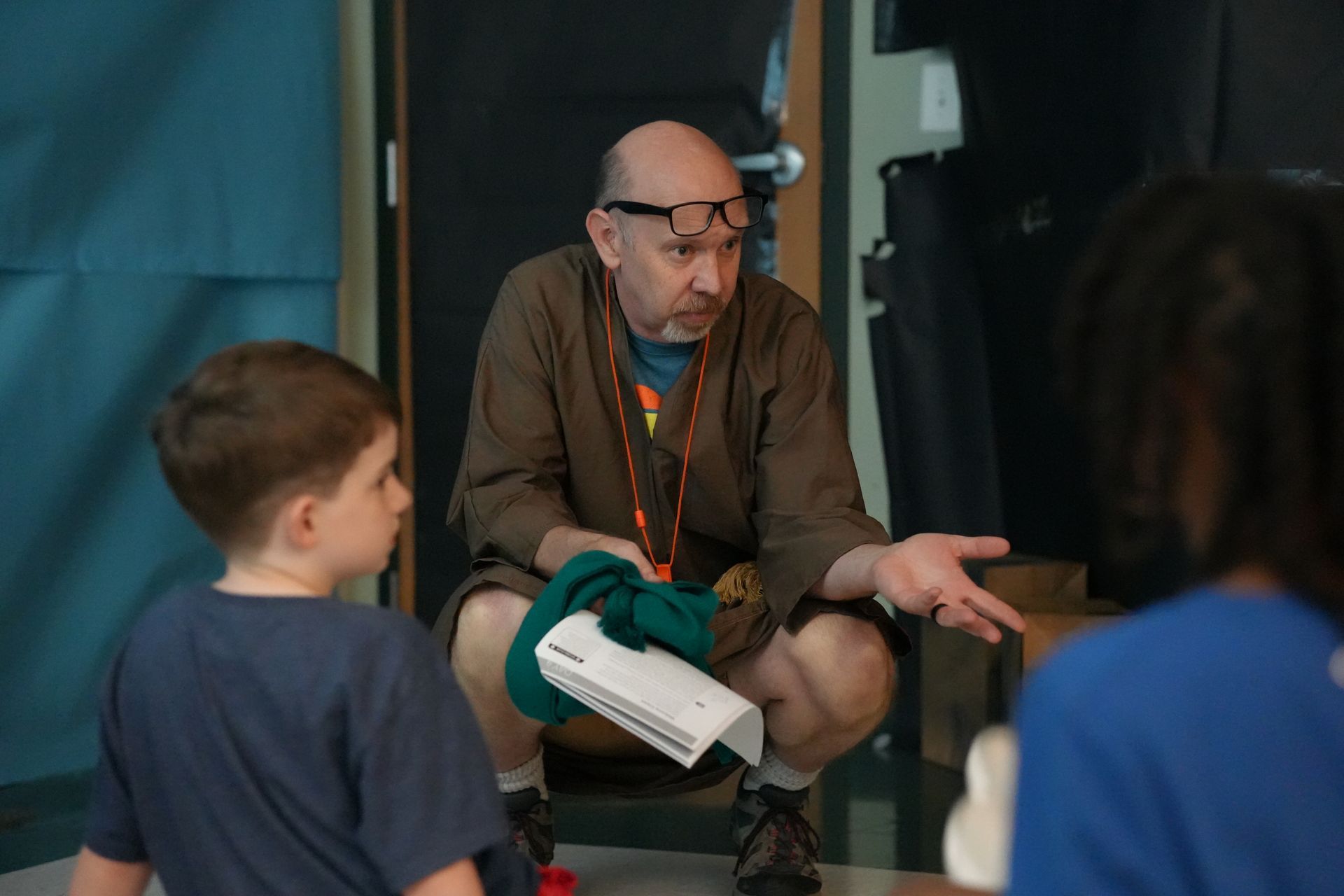 A man wearing glasses is kneeling down and talking to a group of children.